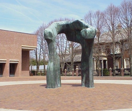 Sculpture. A roughly upside-down U-shaped, abstract bronze sculpture stands in front of a brick building on a brick plaza. The top edge curves towards us, like a C. The patina on the surface is streaked green and grey.