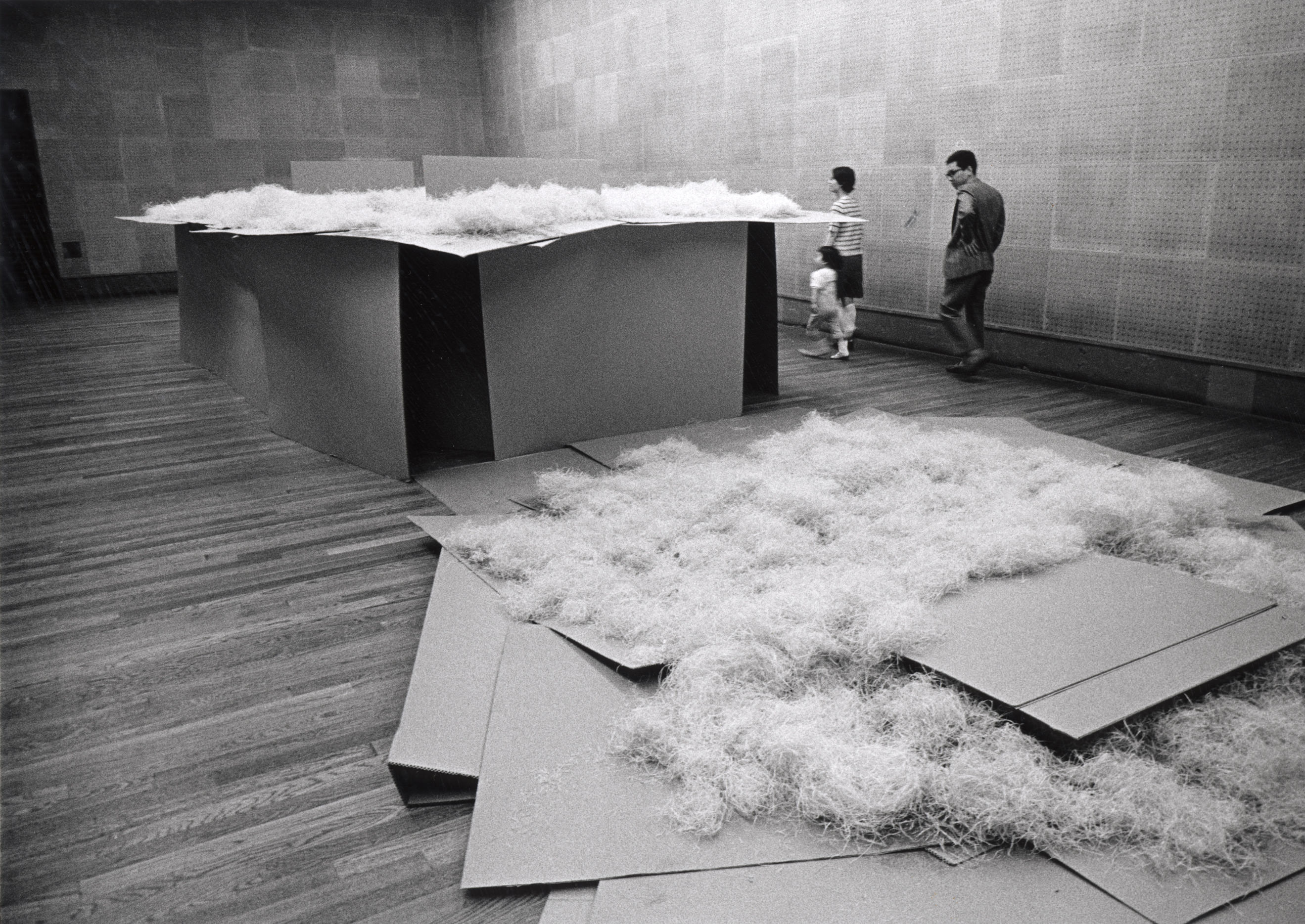 Installation photograph. Black and white photograph shows a table-like sculpture piled with wood shavings. The sides and top are pieces of cardboard stacked like a house of cards. A pile of cardboard and shavings lies on the wood floor, close to us.