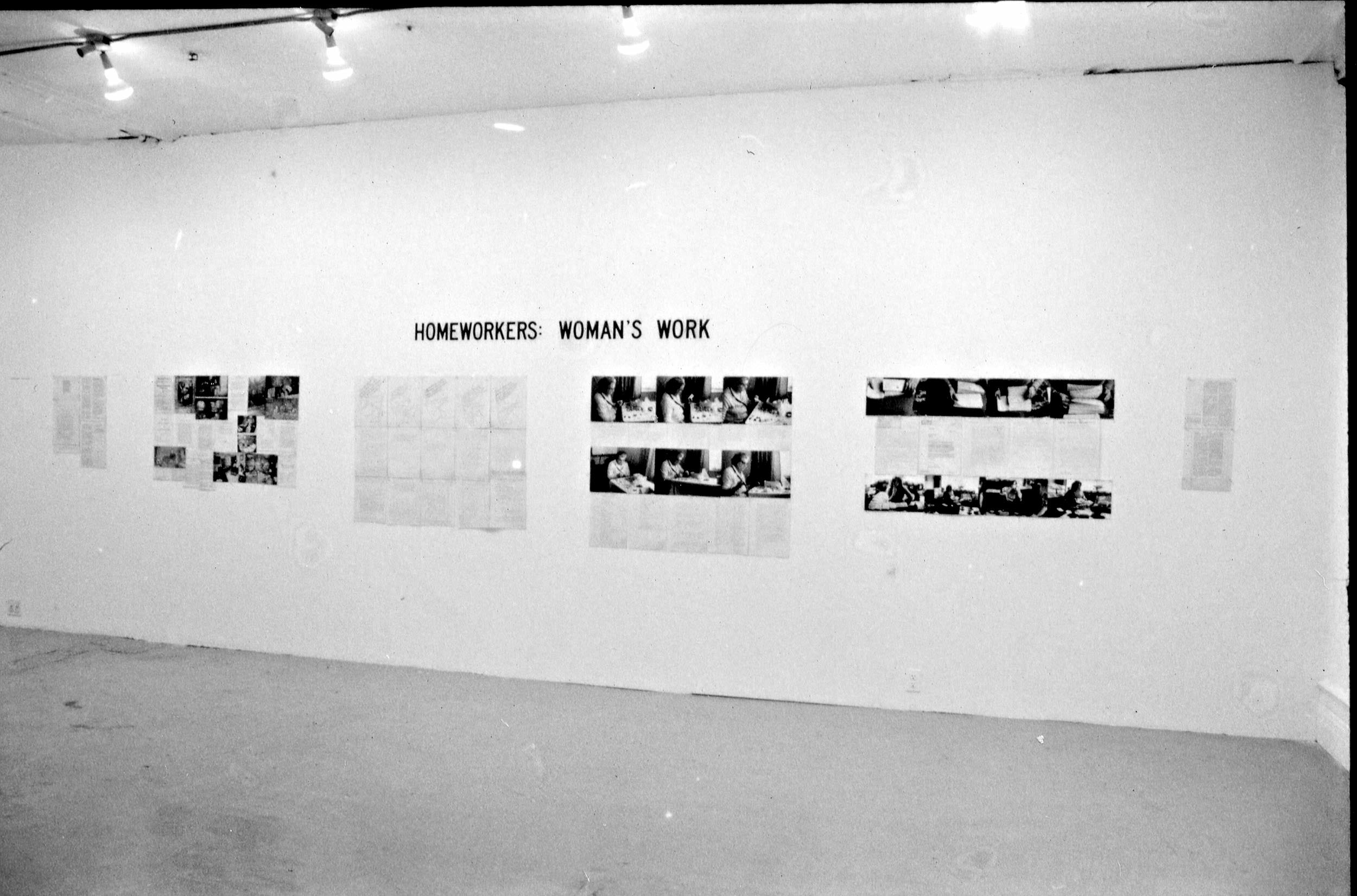 Installation photograph. This black and white photograph shows black and white photographs and printed text hung in five groups on a gallery wall. Above, black text in all caps reads, “Homeworkers: Woman’s Work”.
