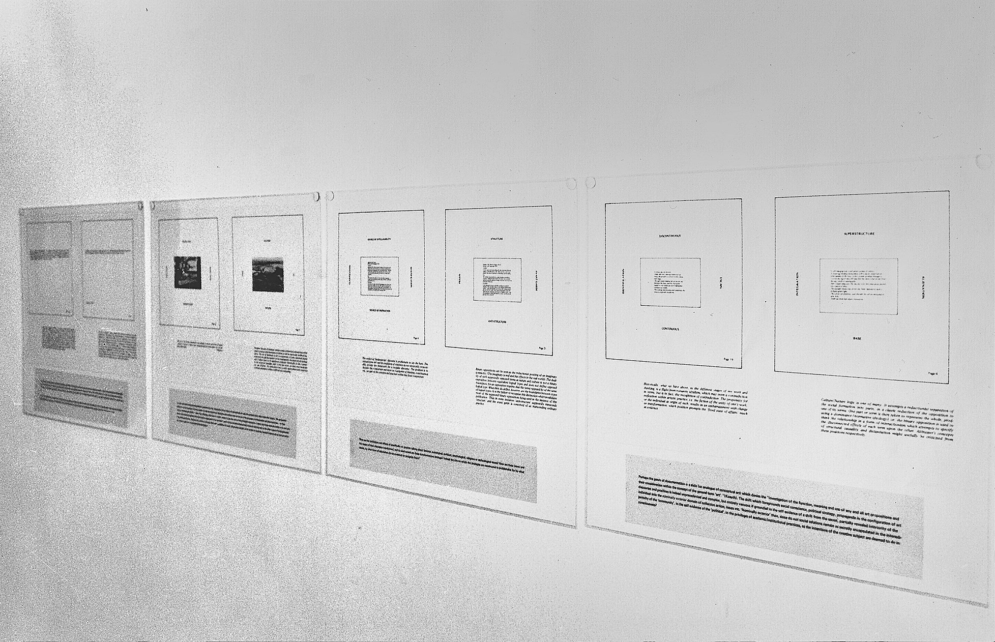 Installation photograph. Black and white photograph of four boards hung on a gallery wall in a row. Each board is printed with schematics like floor plans above blocks of text.