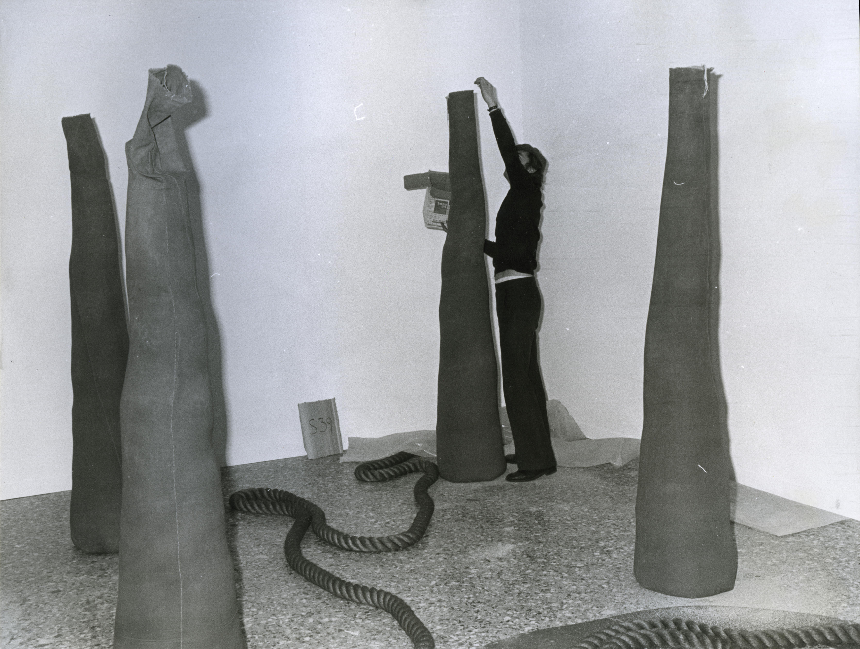 Installation photograph. Four sand-filled, narrow cones stand like sentinels in the corner of a gallery. A man reaches high overhead to drop sand into the cone farthest from us. A thick rope wends across the floor.