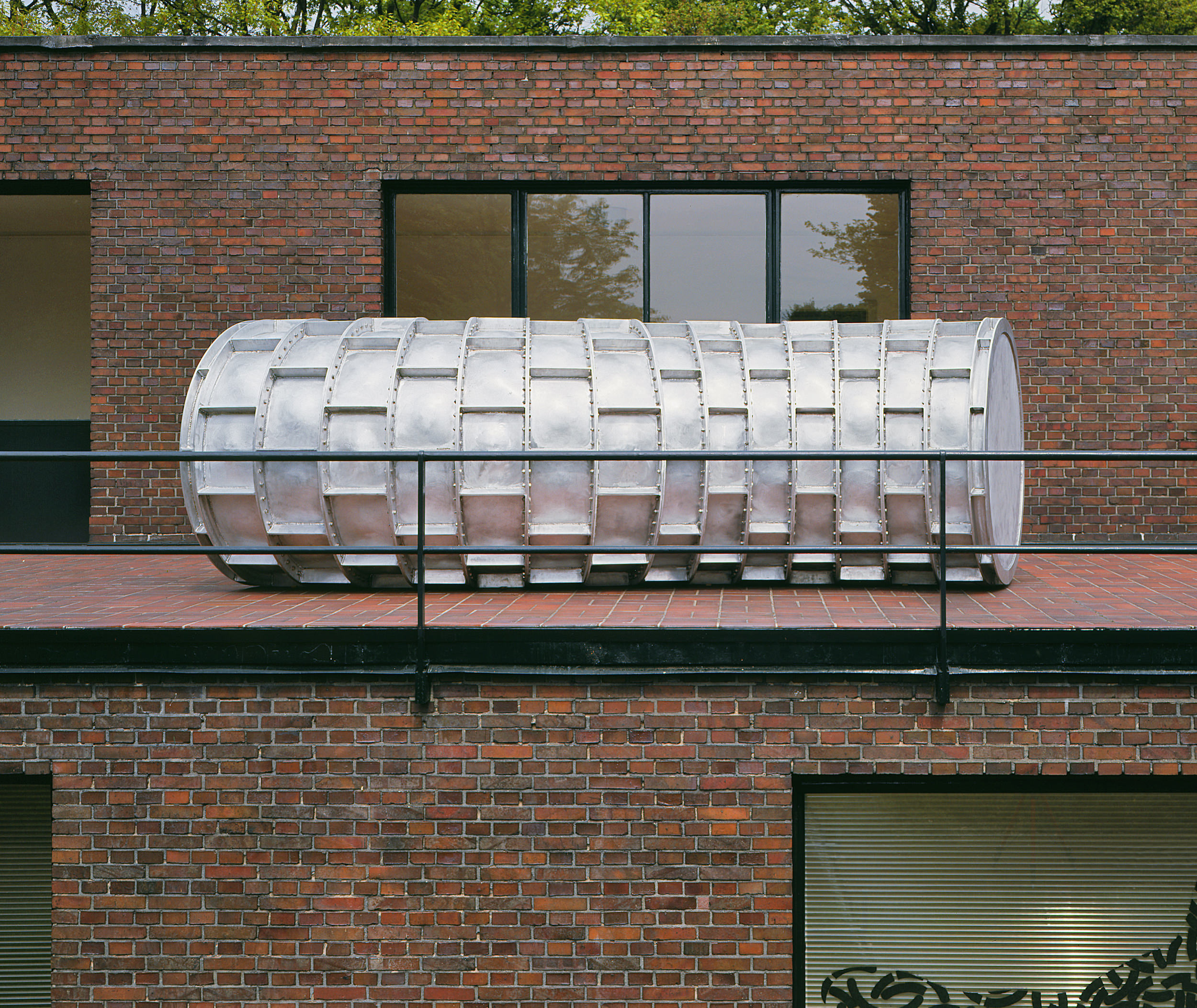 Installation photograph. A long, bright silver pipe made from panels riveted together is on a balcony of a brick building. It spans a double wide window.