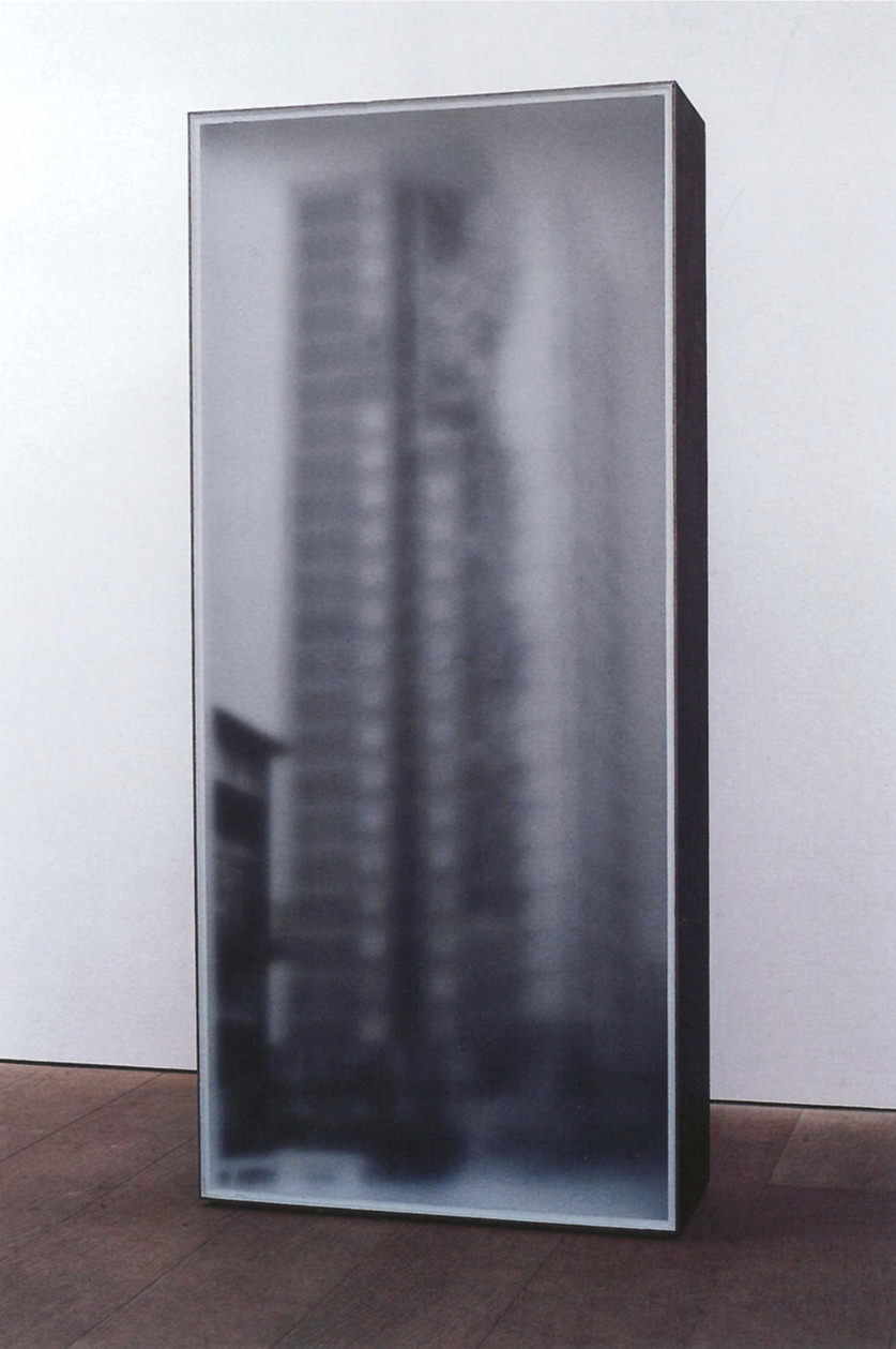 Sculpture. A blurry black and white photograph of a skyscraper is mounted on a tall but shallow, freestanding box in a gallery.