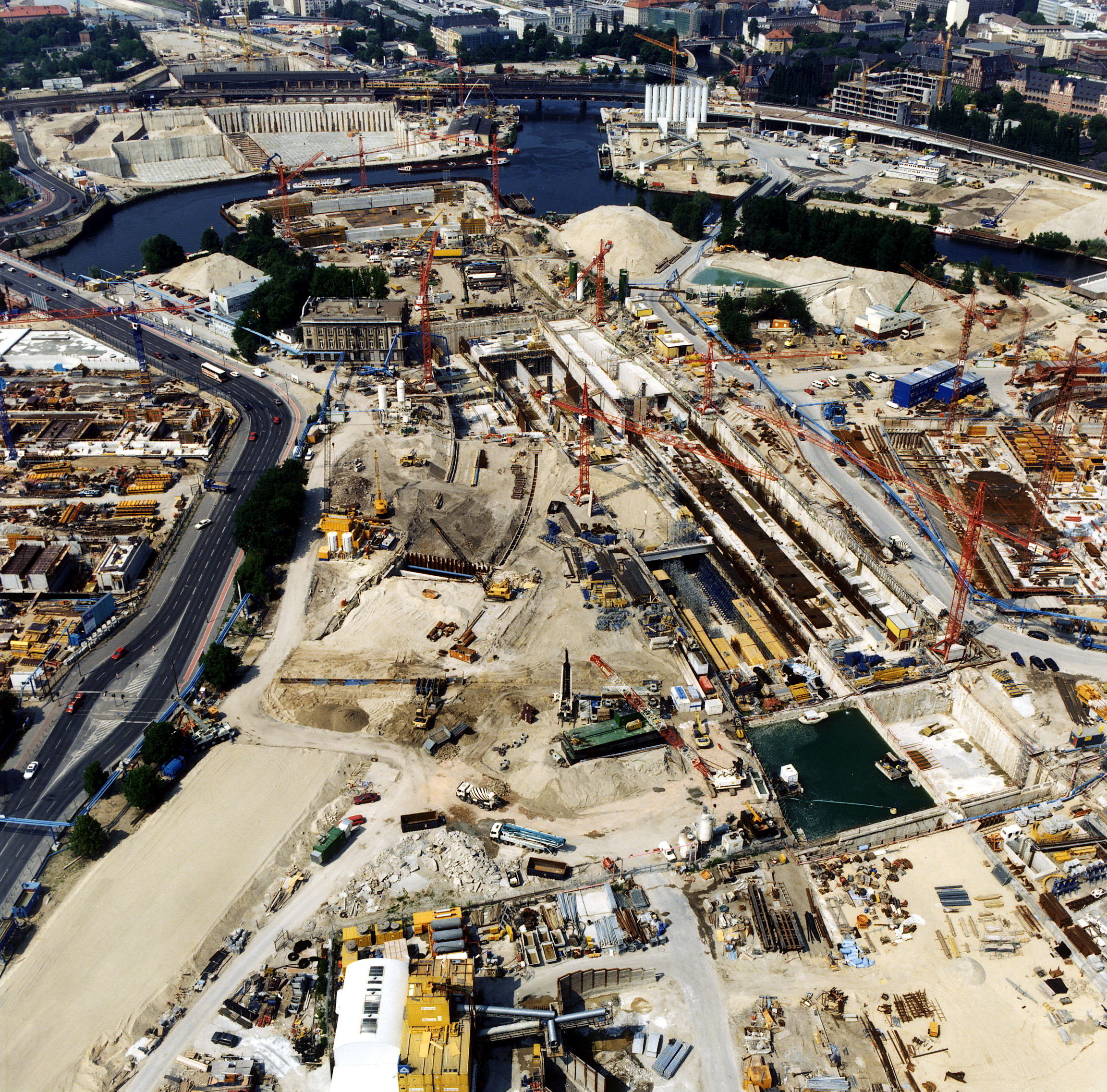 Colour photograph. Aerial view of what appears to be an expansive construction site. A river curves over and through the site, near the top of this photograph. City streets and skyscrapers are visible along the top edge of the image.