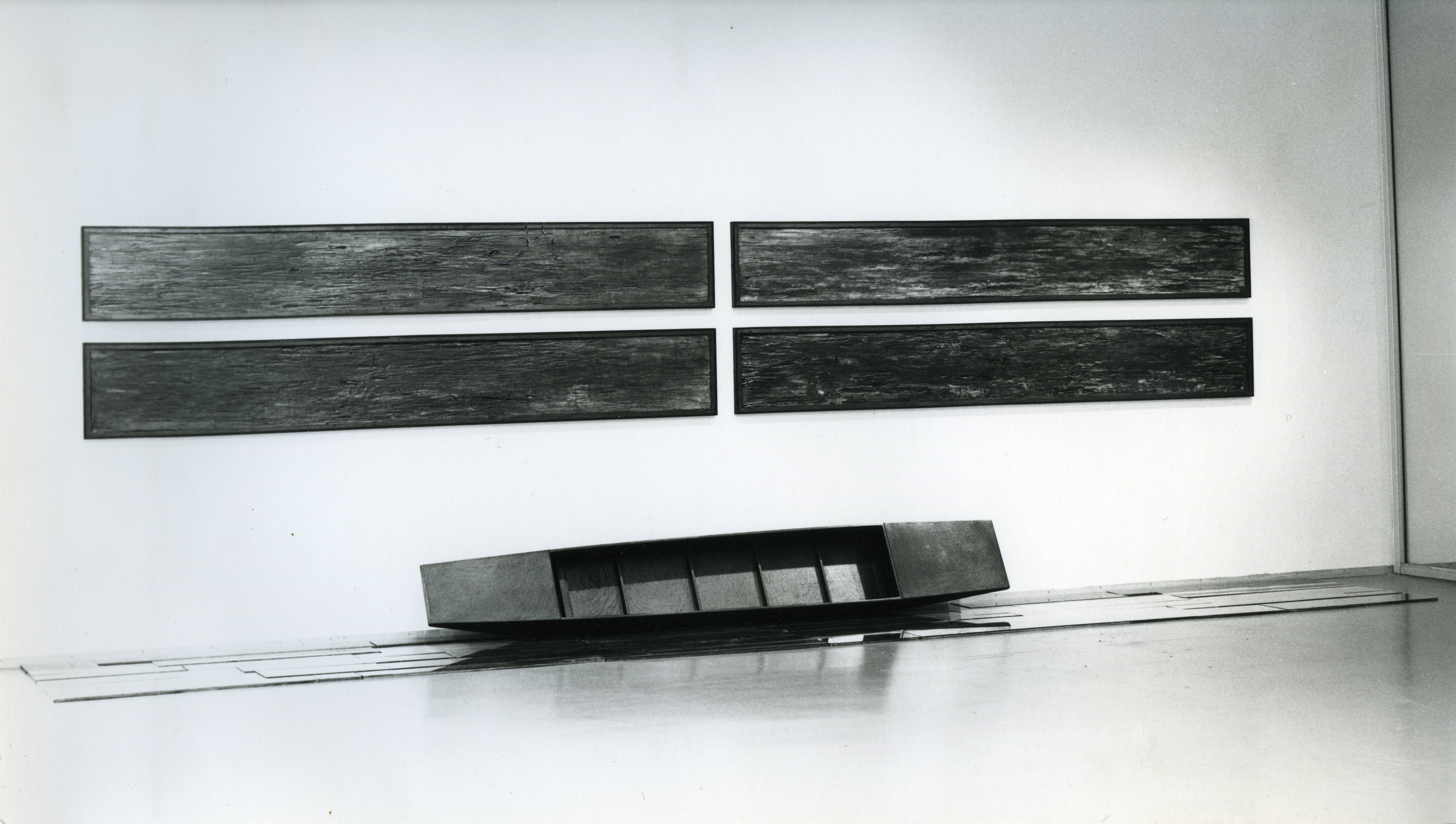 Installation photograph. Black and white photograph of a long, narrow boat propped longways against a gallery wall. Four equally long, textured panels hang above, two by two.