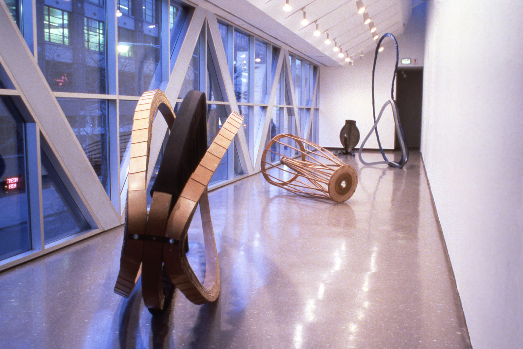 Installation photograph. Four abstract sculptures rest on the floor down a long hallway-like space, which is lined with windows to our left. All use curvilinear shapes made of wood or metal.
