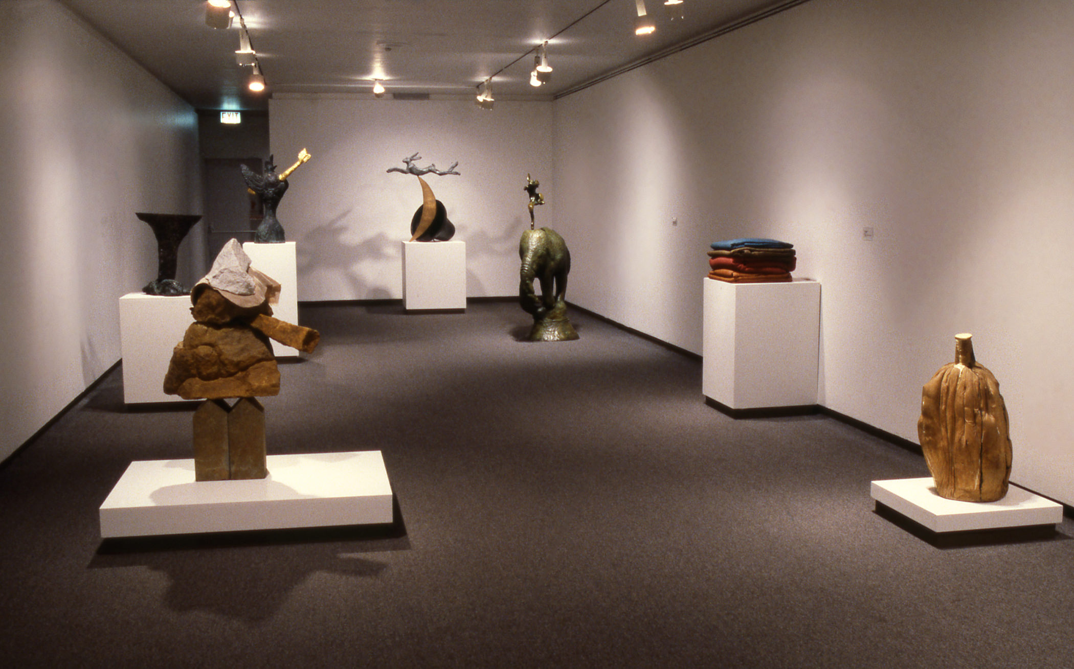 Installation photograph. Seven sculptures are on low or high plinths in a long gallery. Some are abstracted though familiar forms can be found, including a vase, an elephant, and a pile of cloth or clothing.