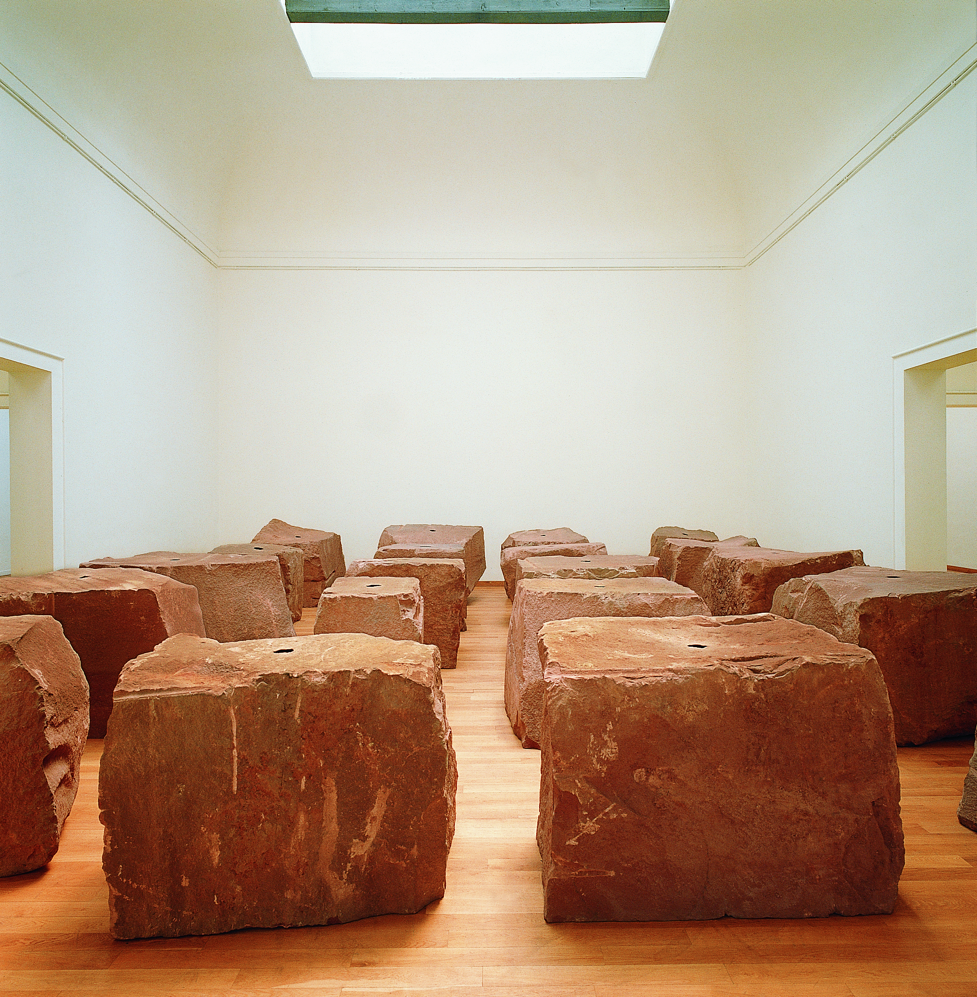 Installation photograph. Twenty roughly cube or rectangular red sandstone cubes sit in rows in a gallery under a skylight. Each has a black circle at the top centre.