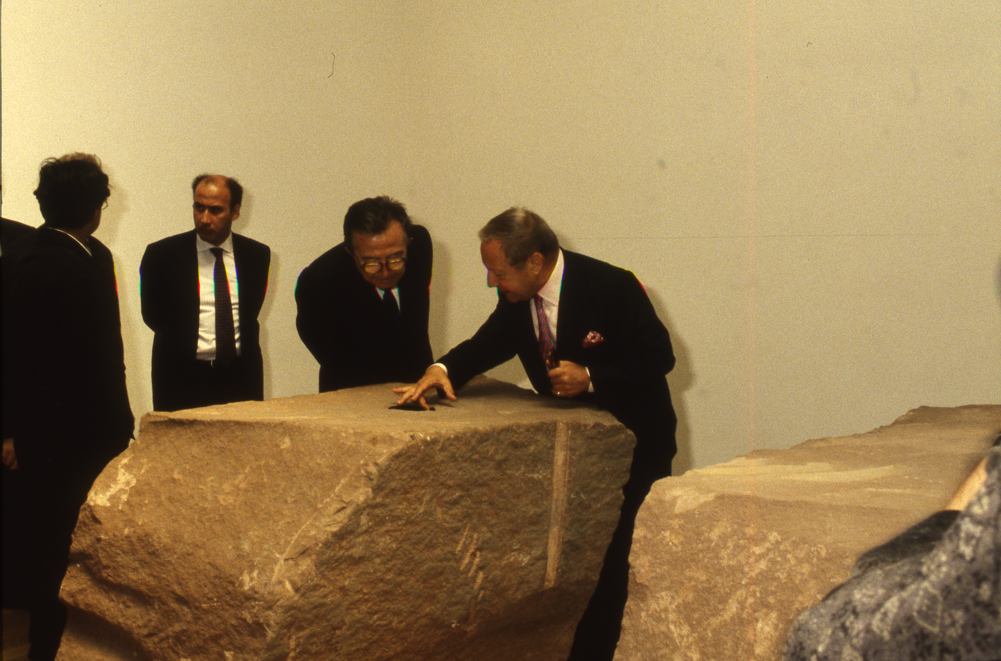 Colour photograph. Five men gather around a roughly rectangular, red sandstone block. One man reaches a middle finger to touch the black circle on the top of the block.