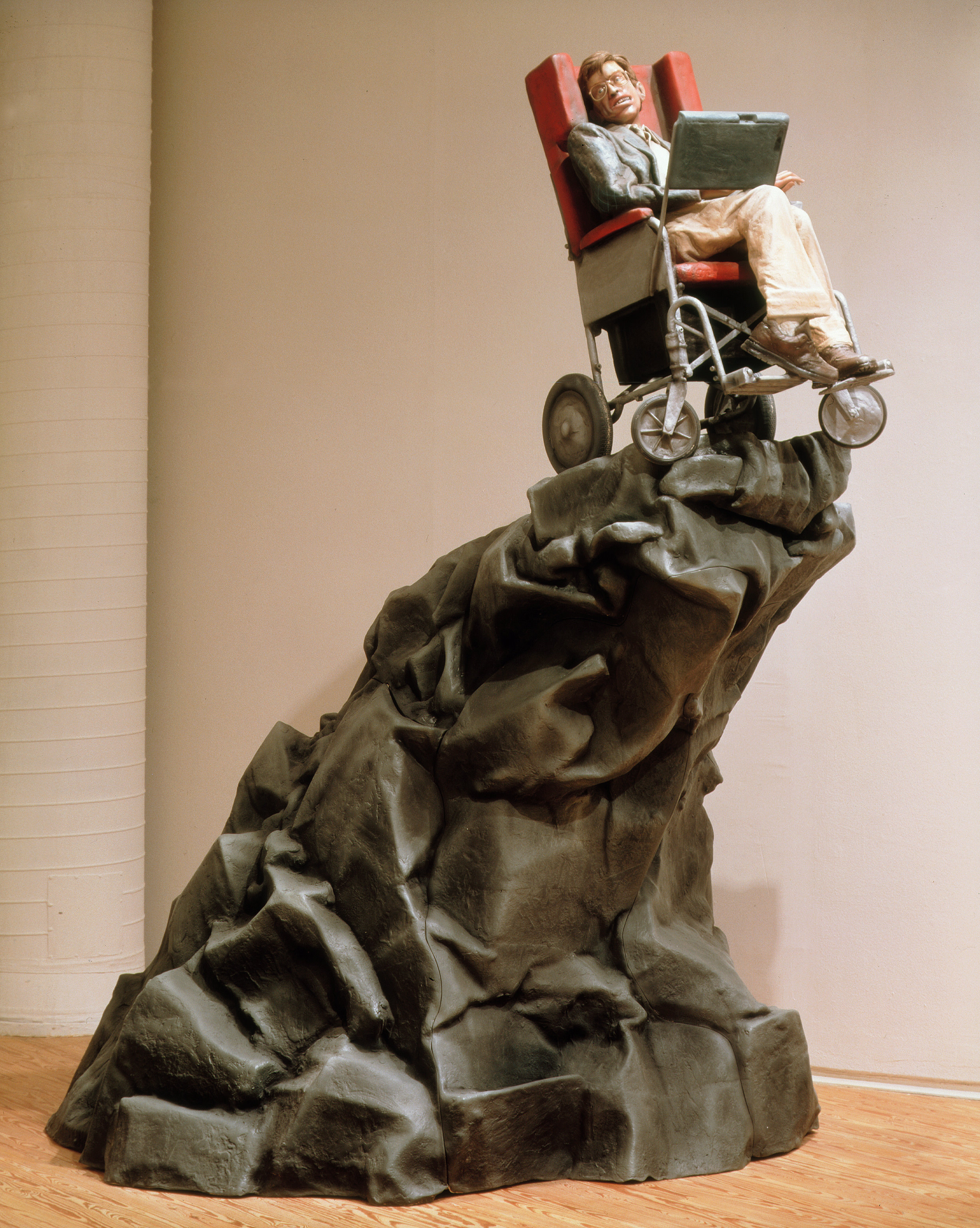 Sculpture. A rocky wave, painted grey, lifts a man in a wheelchair. Stephen Hawking is recognizable from his glasses and wide mouth, as well as his high-sided chair the computer screen over his legs. Hawking and the chair are painted in lifelike colours.