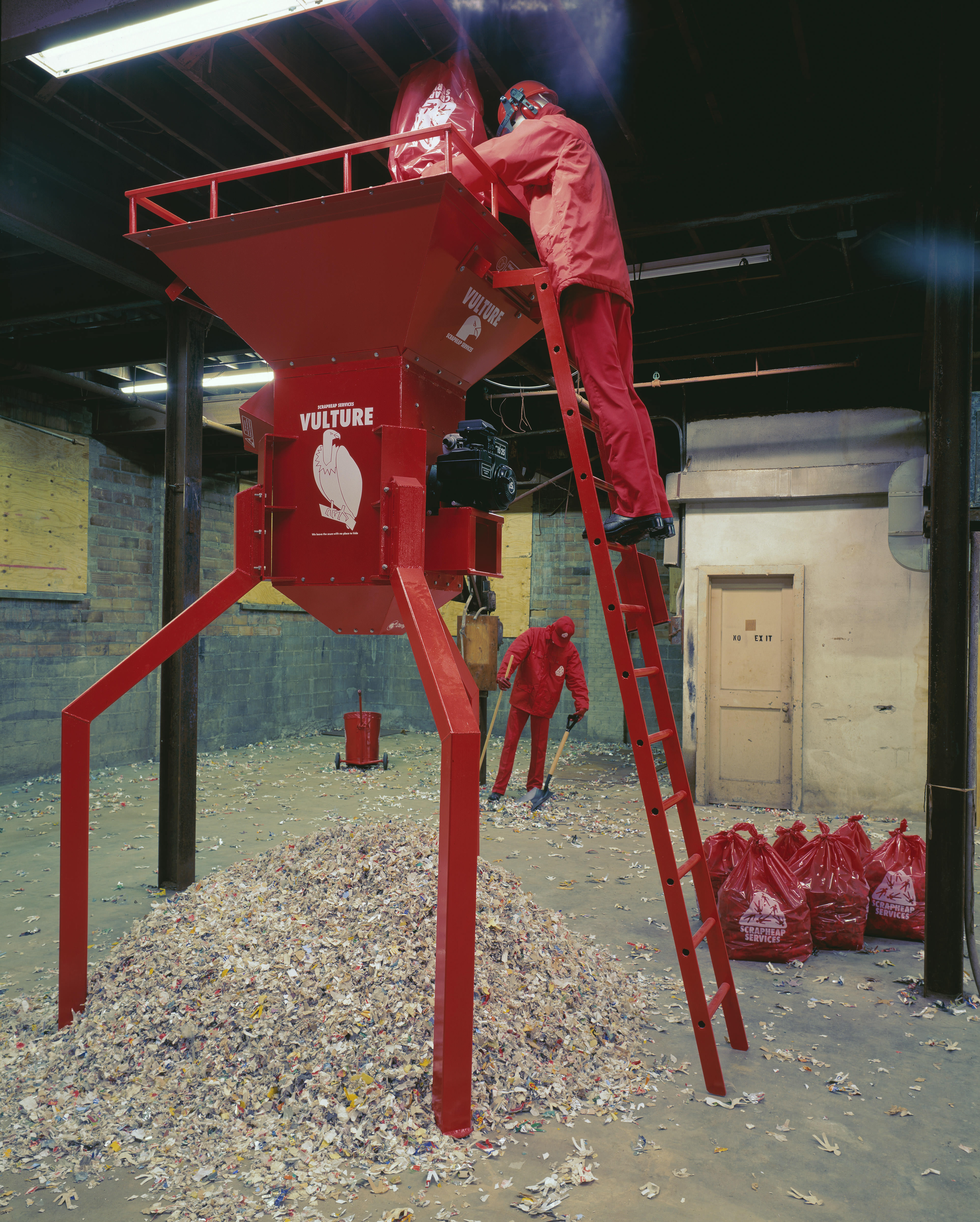 Installation photograph. A mannequin dressed in red stands near the top of a ladder and empties a red bag into a shredder, which is about two storeys high. A mound of shredded material sits under the machine. More red bags are tied to the side, and another red-clad mannequin sweeps in the background.