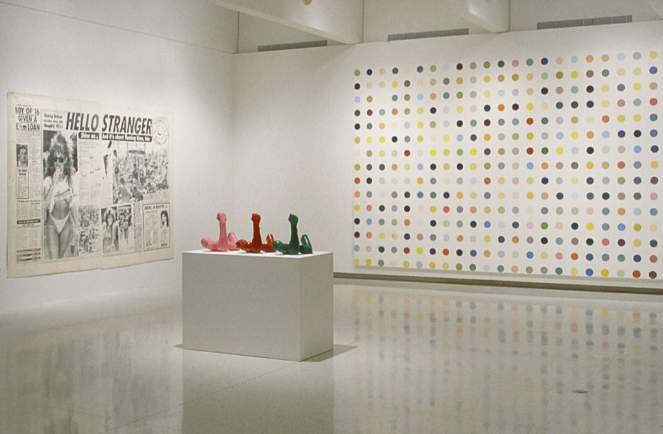 Installation photograph. Two works of art hang on gallery walls and three sculptures share a plinth in the middle of the space. To our left, a dramatically oversized newspaper clipping is titled “Hello Stranger”. A canvas on the wall to our right nearly spans the height of the wall and is painted with a grid of dots, each a single colour.